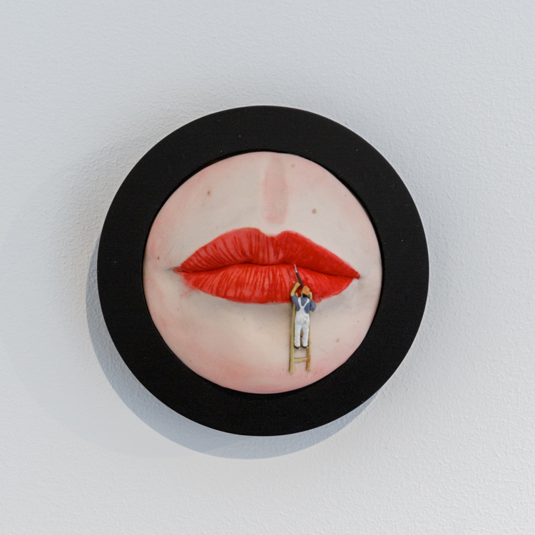 Rond kunstwerk aan witte muure van geschilderde rode lippen in een dikke zwarte cirkel. Tegen de onderste lip staat een mannetje met een kwast op een ladder.